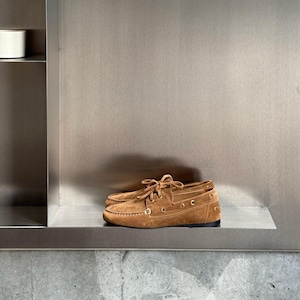 Op de afbeelding: Een paar bruine su&egrave;de bootschoenen met veters, uitgestald op een metalen plank. De schoenen hebben een klassiek ontwerp met zichtbare stiksels en een rubberen zool. De achtergrond is een neutraal metalen oppervlak.
