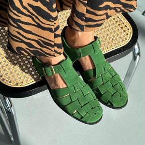 Puede incluir: Sandalias de ante verde brillante con dise&ntilde;o tejido. Las sandalias tienen cierre de hebilla. El calzado es un accesorio vibrante. Se encuentran sobre un fondo claro.