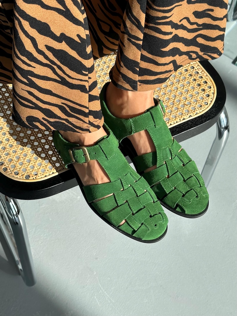 Puede incluir: Sandalias de ante verde brillante con dise&ntilde;o tejido. Las sandalias tienen cierre de hebilla. El calzado es un accesorio vibrante. Se encuentran sobre un fondo claro.