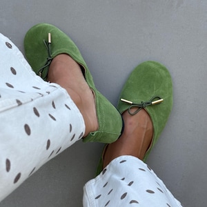 May include: A pair of bright green suede ballet flats featuring a rounded toe, a thin drawstring tie across the top, and small gold-toned metal aglets at the ends of the ties. The shoes are worn with white trousers featuring a brown polka dot pattern.