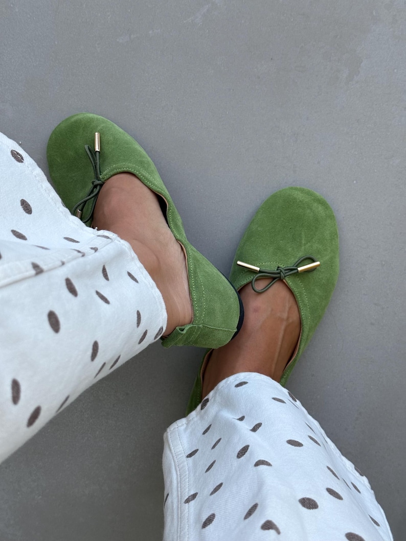 May include: A pair of bright green suede ballet flats featuring a rounded toe, a thin drawstring tie across the top, and small gold-toned metal aglets at the ends of the ties. The shoes are worn with white trousers featuring a brown polka dot pattern.