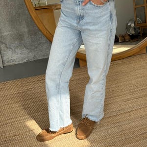 Op de afbeelding: Lichtblauwe jeans met een rafelige zoom, gecombineerd met bruine su&egrave;de mocassins. Een bruine leren riem wordt bij de jeans gedragen. De outfit is casual en comfortabel.
