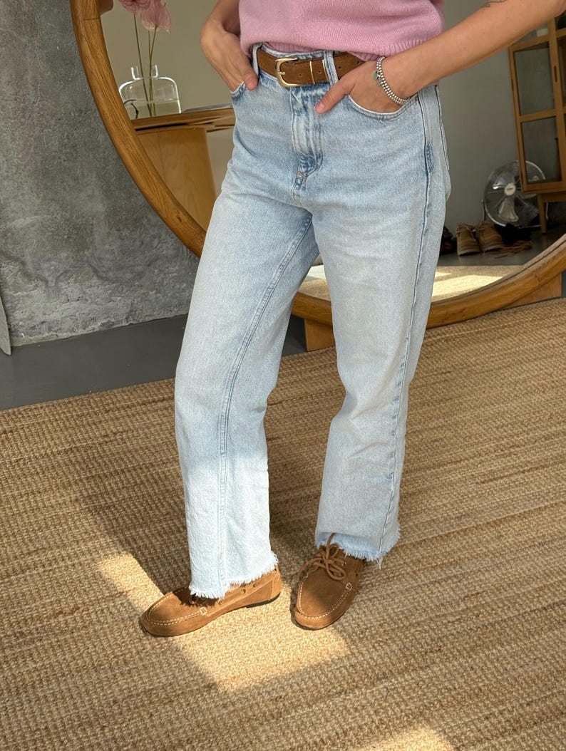 Op de afbeelding: Lichtblauwe jeans met een rafelige zoom, gecombineerd met bruine su&egrave;de mocassins. Een bruine leren riem wordt bij de jeans gedragen. De outfit is casual en comfortabel.