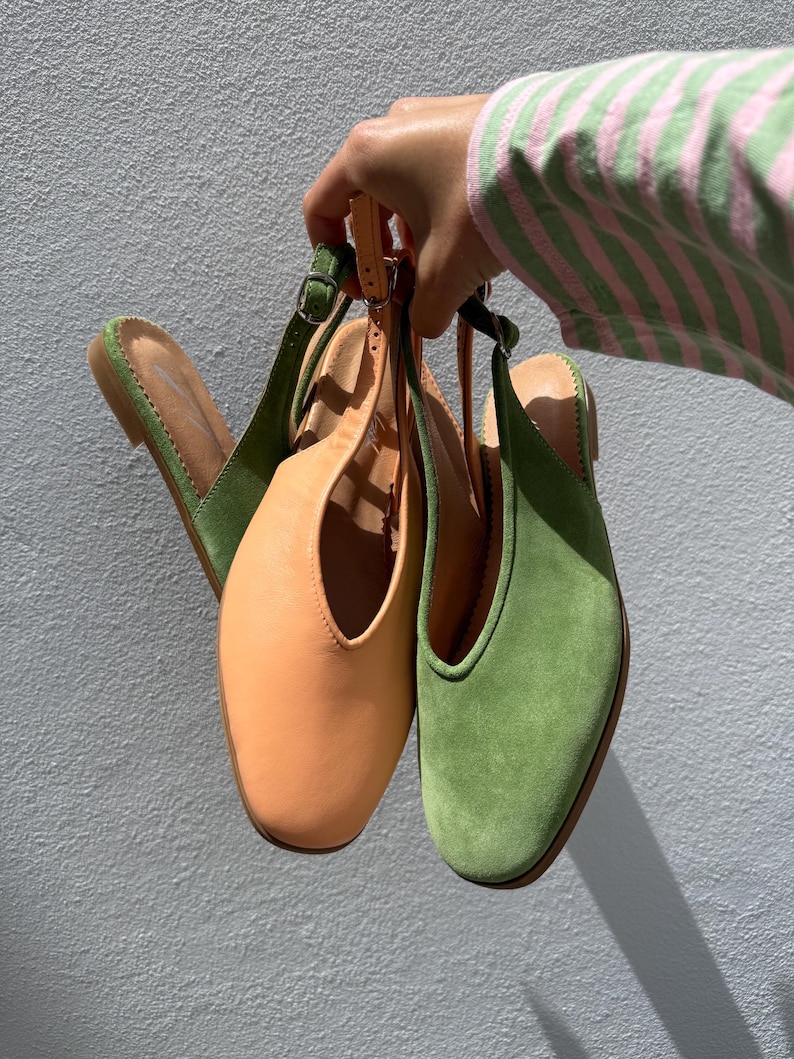 May include: Three pairs of women's slingback shoes in shades of green and peach. The shoes have a square toe design and adjustable straps. The shoes are held up against a light gray wall.