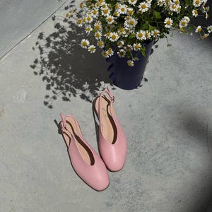 May include: A pair of light pink slingback flats with a rounded toe, resting on a concrete surface. A dark blue bucket filled with white and yellow daisies is in the background, casting a shadow.