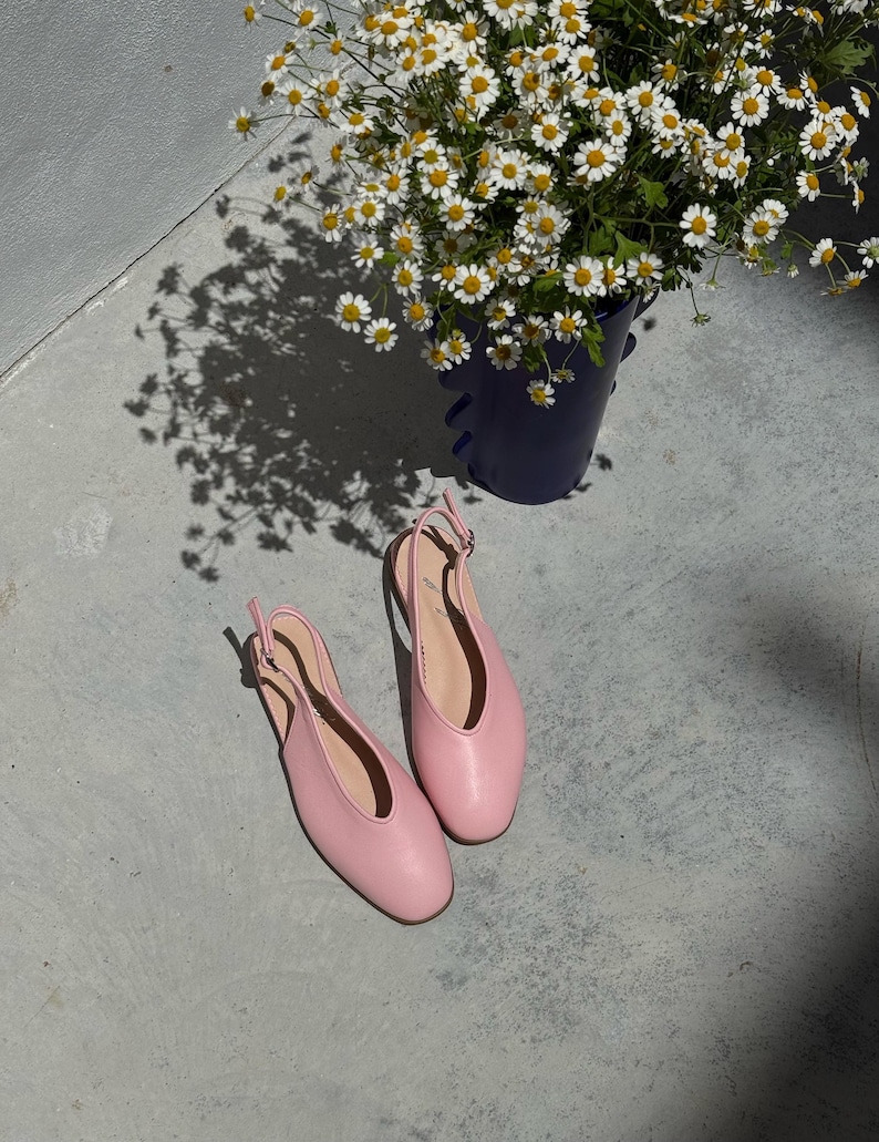 May include: A pair of light pink slingback flats with a rounded toe, resting on a concrete surface. A dark blue bucket filled with white and yellow daisies is in the background, casting a shadow.