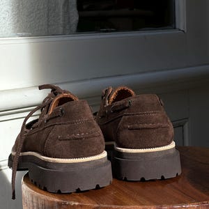 May include: A pair of brown suede boat shoes with thick, dark brown rubber soles. The shoes have laces and a classic boat shoe design, with visible stitching and a light-colored trim around the sole. The shoes are placed on a wooden surface.