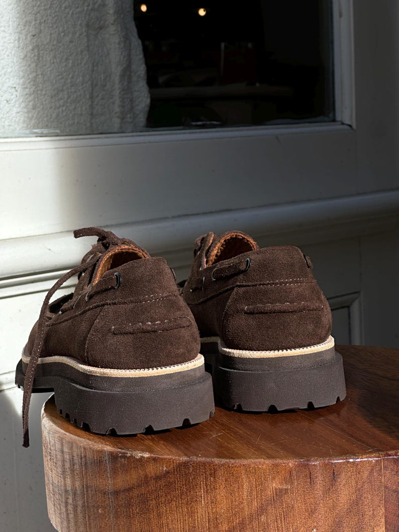 May include: A pair of brown suede boat shoes with thick, dark brown rubber soles. The shoes have laces and a classic boat shoe design, with visible stitching and a light-colored trim around the sole. The shoes are placed on a wooden surface.