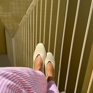 May include: A pair of cream-colored, pointed-toe flats. The shoes are resting on a white surface, with a pink and white striped garment visible. The background features a yellow wall and white railing.