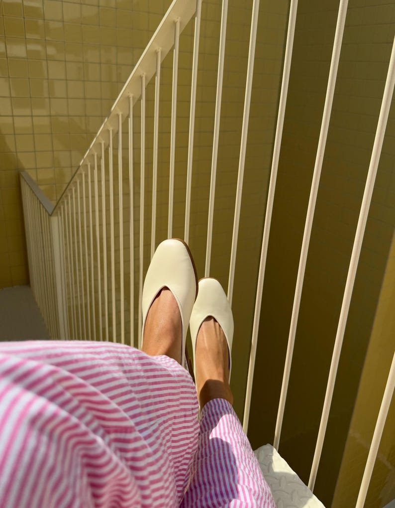 May include: A pair of cream-colored, pointed-toe flats. The shoes are resting on a white surface, with a pink and white striped garment visible. The background features a yellow wall and white railing.