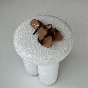 May include: A pair of brown sandals with dark brown straps and gold-colored buckles rests on a white, textured stool. The sandals have a minimalist design, with a single strap across the toes and an ankle strap.