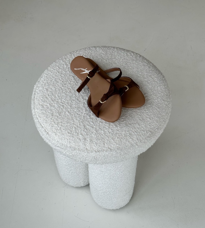 May include: A pair of brown sandals with dark brown straps and gold-colored buckles rests on a white, textured stool. The sandals have a minimalist design, with a single strap across the toes and an ankle strap.
