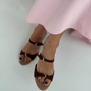 May include: Brown suede sandals with ankle straps and small gold buckles. The sandals have a bow detail on the toe strap and a tan sole. The image also shows a pink dress.