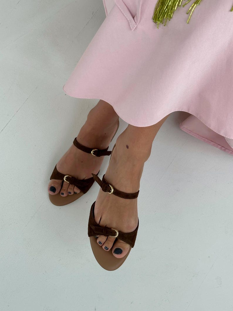 May include: Brown suede sandals with ankle straps and small gold buckles. The sandals have a bow detail on the toe strap and a tan sole. The image also shows a pink dress.