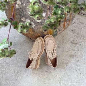 May include: A pair of beige suede shoes with a rounded toe and thick, light-brown soles. The shoes have laces and a classic moccasin design. The image is taken from a high angle, with the shoes resting on a concrete surface.