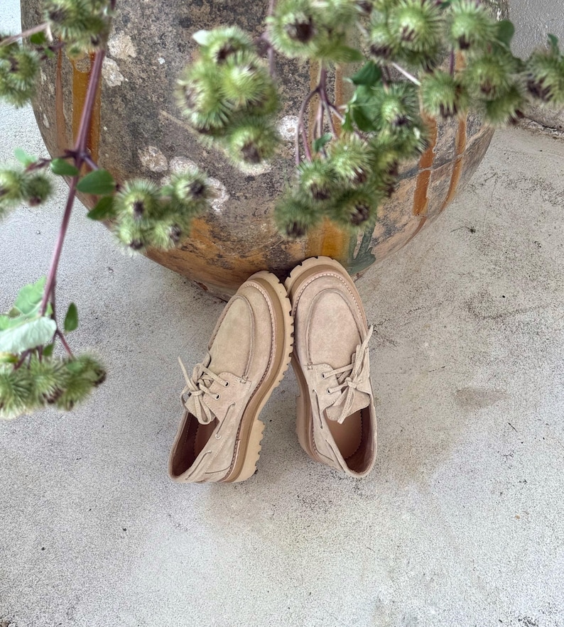May include: A pair of beige suede shoes with a rounded toe and thick, light-brown soles. The shoes have laces and a classic moccasin design. The image is taken from a high angle, with the shoes resting on a concrete surface.