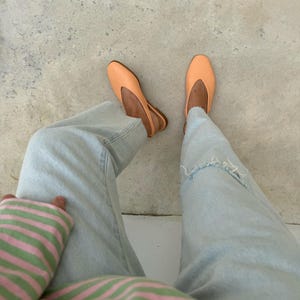 May include: A pair of light orange ballet flats with a slingback design. The shoes are worn with light blue denim jeans and a striped pink and green top. The image is taken from a top-down perspective.
