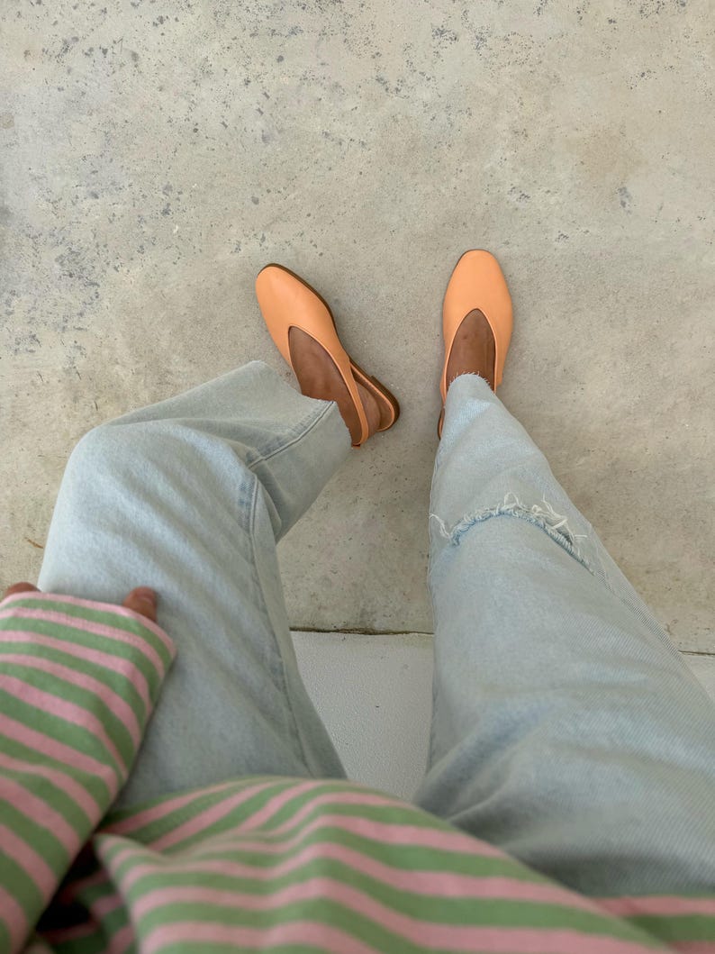 May include: A pair of light orange ballet flats with a slingback design. The shoes are worn with light blue denim jeans and a striped pink and green top. The image is taken from a top-down perspective.