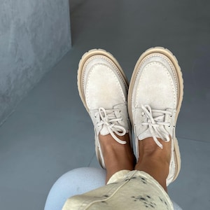 May include: A pair of off-white suede boat shoes with beige soles and laces. The shoes have a classic design with visible stitching and a rounded toe. The image shows the shoes from a top-down perspective.