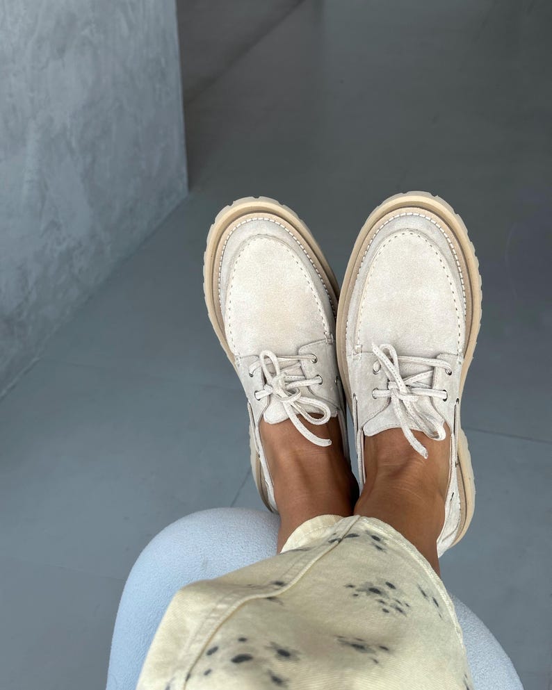 May include: A pair of off-white suede boat shoes with beige soles and laces. The shoes have a classic design with visible stitching and a rounded toe. The image shows the shoes from a top-down perspective.