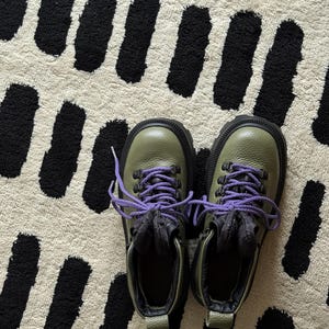 May include: A pair of olive green hiking boots with black soles and purple laces. The boots are on a cream-colored rug with a black abstract pattern.