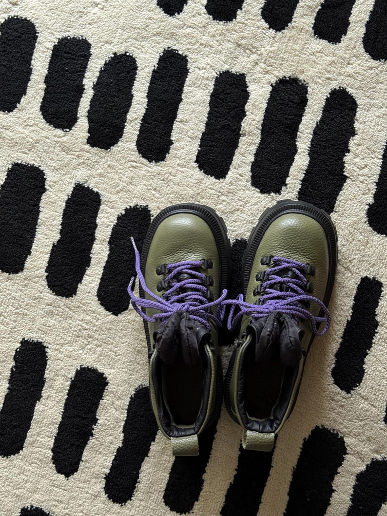 May include: A pair of olive green hiking boots with black soles and purple laces. The boots are on a cream-colored rug with a black abstract pattern.