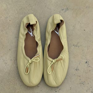 May include: A pair of pale yellow ballet flats featuring a soft, gathered leather upper, a rounded toe, and a decorative drawstring tie at the front with metallic gold-toned aglets. The interior footbed is brown and displays a white script logo.