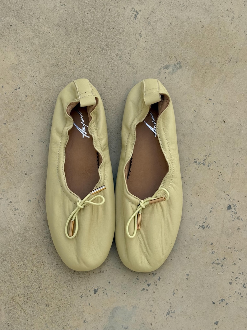 May include: A pair of pale yellow ballet flats featuring a soft, gathered leather upper, a rounded toe, and a decorative drawstring tie at the front with metallic gold-toned aglets. The interior footbed is brown and displays a white script logo.