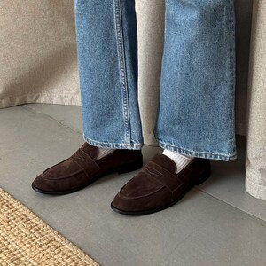 May include: A pair of brown suede loafers with a strap across the top. The shoes have a rounded toe and a black sole. The person is wearing blue jeans and white socks.