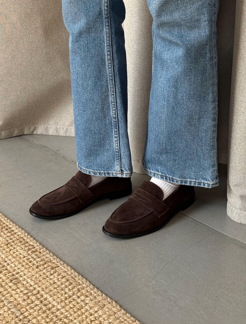 May include: A pair of brown suede loafers with a strap across the top. The shoes have a rounded toe and a black sole. The person is wearing blue jeans and white socks.