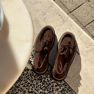 May include: A pair of brown suede boat shoes with white stitching and laces. The shoes have a classic design with a rounded toe and a rubber sole. The shoes are displayed on a patterned tile surface.