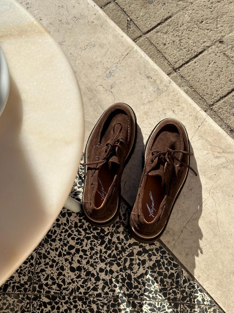 May include: A pair of brown suede boat shoes with white stitching and laces. The shoes have a classic design with a rounded toe and a rubber sole. The shoes are displayed on a patterned tile surface.