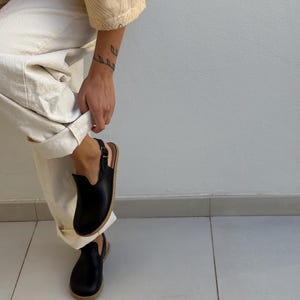 May include: Black leather clogs with a buckle fastening and a light brown sole. The clogs have a rounded toe and a low heel. The person is wearing off-white trousers and a beige top.