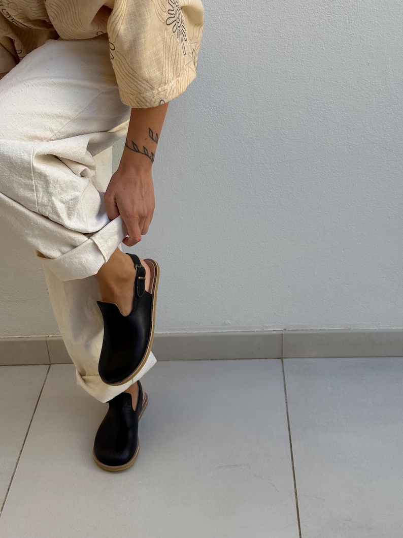 May include: Black leather clogs with a buckle fastening and a light brown sole. The clogs have a rounded toe and a low heel. The person is wearing off-white trousers and a beige top.