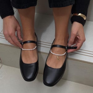 May include: Black leather flats with a pearl ankle strap and a small gold charm. The shoes have a classic design with a rounded toe and a buckle closure. The flats are being worn.