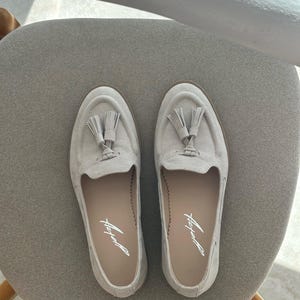 May include: A pair of light grey suede loafers with tassel detailing. The shoes have a rounded toe and a low heel. The brand name is printed in script on the insole. The shoes are displayed on a grey chair.