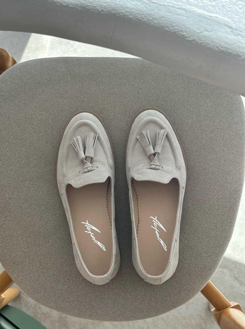 May include: A pair of light grey suede loafers with tassel detailing. The shoes have a rounded toe and a low heel. The brand name is printed in script on the insole. The shoes are displayed on a grey chair.