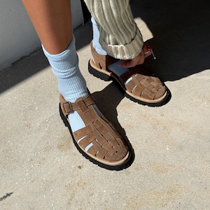 Puede incluir: Sandalias de ante marr&oacute;n con dise&ntilde;o tejido. Las sandalias tienen suela negra y ribete claro. La persona lleva calcetines azul claro y un su&eacute;ter claro.