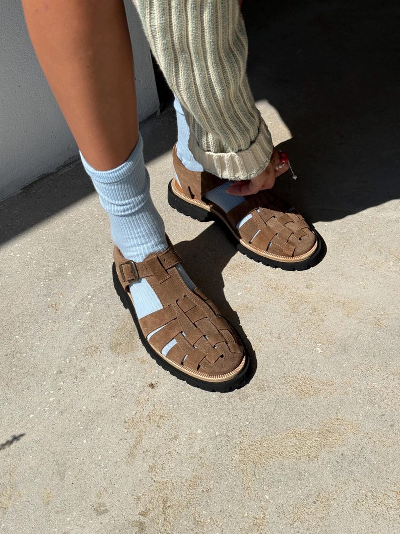 Puede incluir: Sandalias de ante marr&oacute;n con dise&ntilde;o tejido. Las sandalias tienen suela negra y ribete claro. La persona lleva calcetines azul claro y un su&eacute;ter claro.