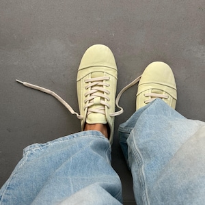 Peut inclure: Vue de dessus de baskets jaune pâle avec des lacets crème. Les baskets sont portées avec un jean en denim bleu clair. Les chaussures sont sur une surface grise.