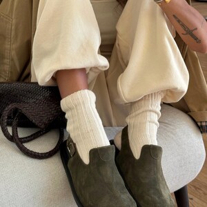 May include: Olive green suede clogs with black soles. The clogs are paired with cream-colored ribbed socks and beige sweatpants. A brown leather bag and a tan jacket complete the casual outfit.