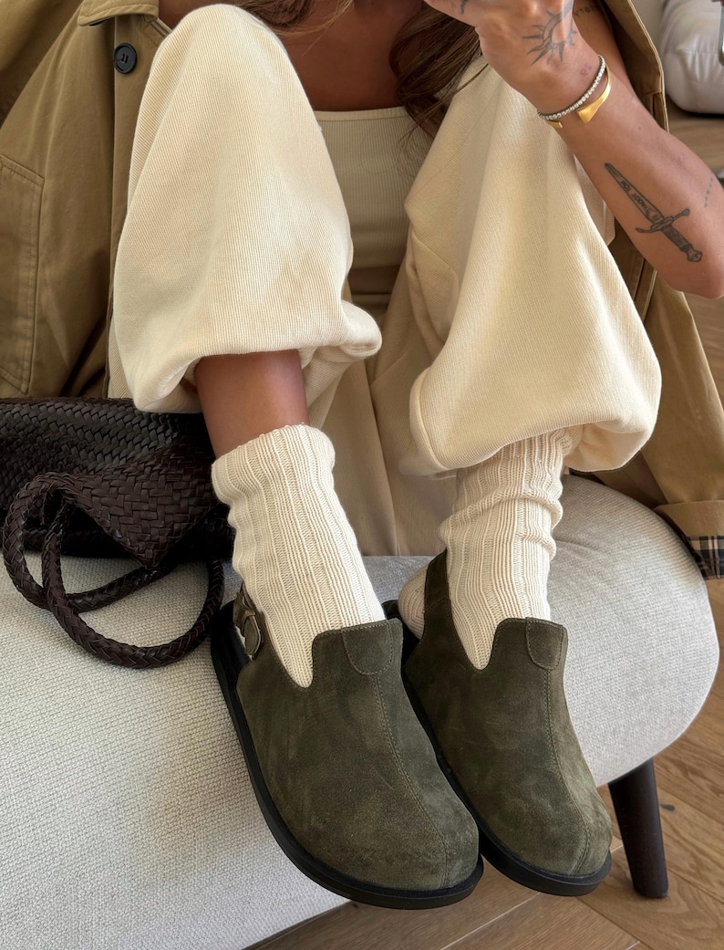May include: Olive green suede clogs with black soles. The clogs are paired with cream-colored ribbed socks and beige sweatpants. A brown leather bag and a tan jacket complete the casual outfit.