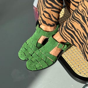 Puede incluir: Un par de sandalias de ante verde con un dise&ntilde;o tejido. Las sandalias tienen una correa con cierre de hebilla. La persona que lleva las sandalias tambi&eacute;n lleva pantalones con estampado de cebra marr&oacute;n y negro.