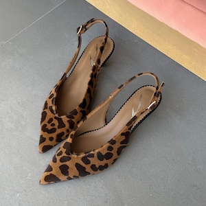 Op de afbeelding: Een paar slingback flats met een luipaardprint. De schoenen hebben een spitse neus en een kleine hak. De enkelband sluit met een kleine gesp. De binnenkant van de schoen is lichtbruin.