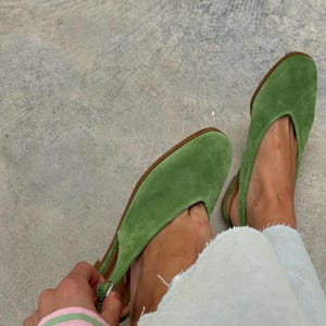 May include: A pair of green suede slingback flats with a pointed toe. The shoes have a brown sole and a strap around the heel. The person is wearing light blue jeans. The background is a gray concrete surface.
