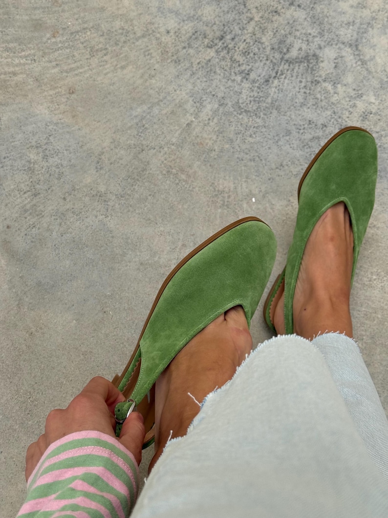 May include: A pair of green suede slingback flats with a pointed toe. The shoes have a brown sole and a strap around the heel. The person is wearing light blue jeans. The background is a gray concrete surface.