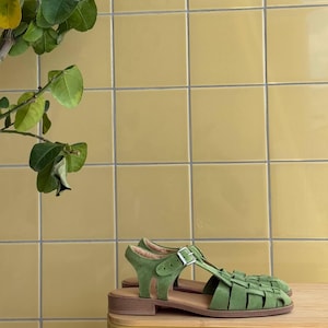 K&ouml;nnte beinhalten: Gr&uuml;ne Wildleder-Sandalen mit geflochtenem Design und Schnallenverschluss. Die Sandalen haben einen niedrigen Absatz und werden auf einer Holzoberfl&auml;che vor einem gelben Fliesen Hintergrund pr&auml;sentiert. Eine gr&uuml;ne Pflanze befindet sich in der oberen linken Ecke.