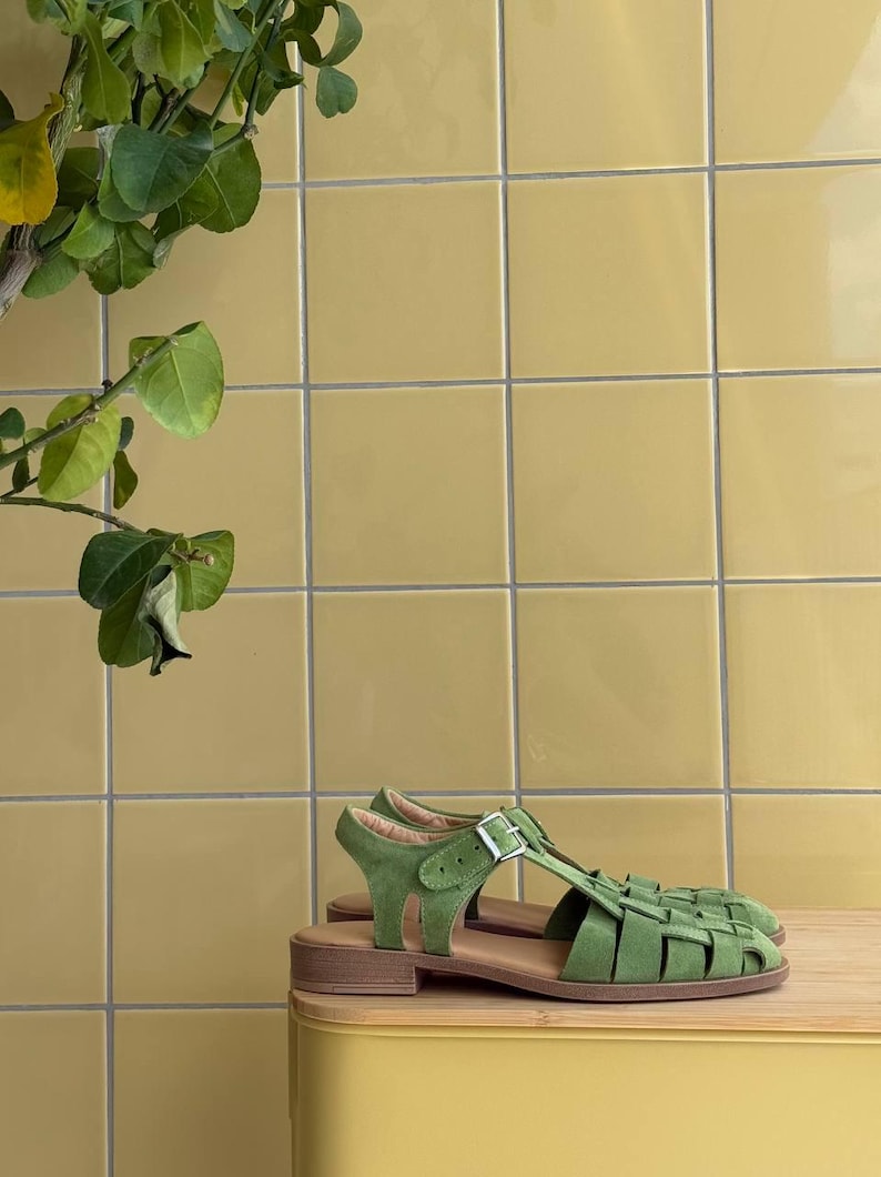 K&ouml;nnte beinhalten: Gr&uuml;ne Wildleder-Sandalen mit geflochtenem Design und Schnallenverschluss. Die Sandalen haben einen niedrigen Absatz und werden auf einer Holzoberfl&auml;che vor einem gelben Fliesen Hintergrund pr&auml;sentiert. Eine gr&uuml;ne Pflanze befindet sich in der oberen linken Ecke.