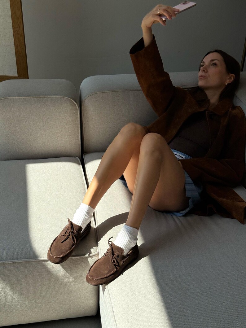 Op de afbeelding: Bruine su&egrave;de bootschoenen met veters, gecombineerd met witte sokken. De schoenen worden gedragen met een bruine jas en shorts. De persoon zit op een lichtgekleurde bank.