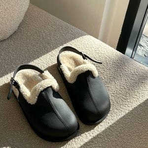 Pode incluir: Um par de tamancos de couro preto com um forro interior cremoso e fofo. Os tamancos têm uma tira à volta do calcanhar e estão sobre uma superfície de tons neutros. Os sapatos parecem novos e por usar.